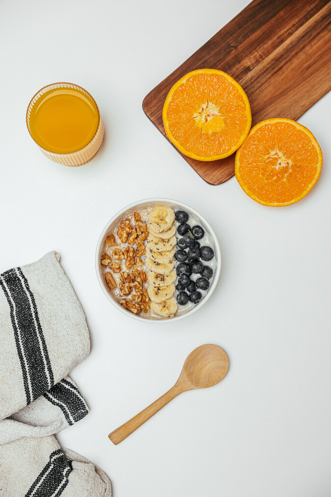En skål med yoghurt eller gröt toppad med banan, valnötter och blåbär, med apelsin, apelsinjuice och en träslev på ett vitt bord